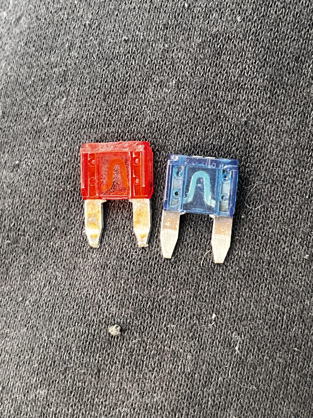 Comparación visual entre un fusible de auto quemado, de color rojo con el filamento roto, y un fusible nuevo de color azul con el filamento intacto.
